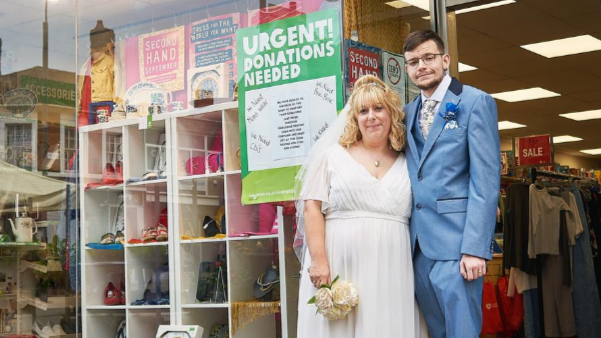 Susan and James Boyles on their wedding day. She is wearing a white wedding dress and he is wearing a blue suit. They are in front of an Oxfam shop. She is holding some flowers.
