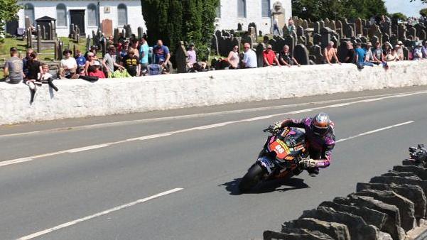 Craig Szczypek leaning over as he travels on the circuit which has stone walls on either side and spectators looking on.