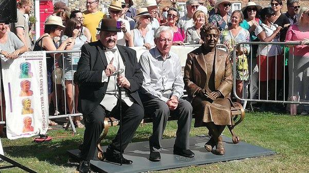 Poirot impersonator sitting next to Agatha Christie's grandson on the bench next to her statue. A crowd of visitors cheer from behind. It's a sunny day.