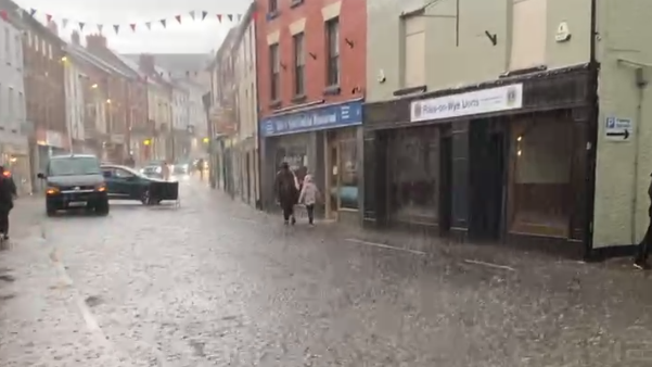 A street with a row of shops and bunting overhead with heavy rain falling and two cars and two pedestrians in the distance