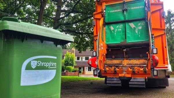 A Shropshire Council green bin on the left, with an orange bin lorry on the right hand side