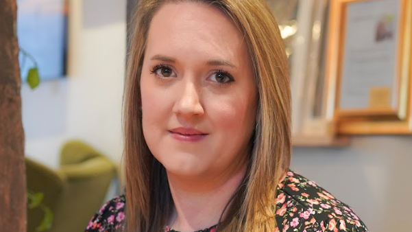A head and shoulders shot of Helen Blythe. She wears a black dress with pink flowers on it. Her brown straight hair is shoulder length and straight.