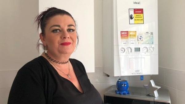 Nikki Byrne stands in her kitchen next to the wall-mounted boiler which has a danger sign stuck on the front of it, bearing the words "do not use". She has dark hair tied back and wears a black top.