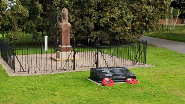 Two war memorials on a small patch of grass. One stands a few feet tall and is surrounded by a fence. The other is a black plinth with two poppy wreaths leaning against it.