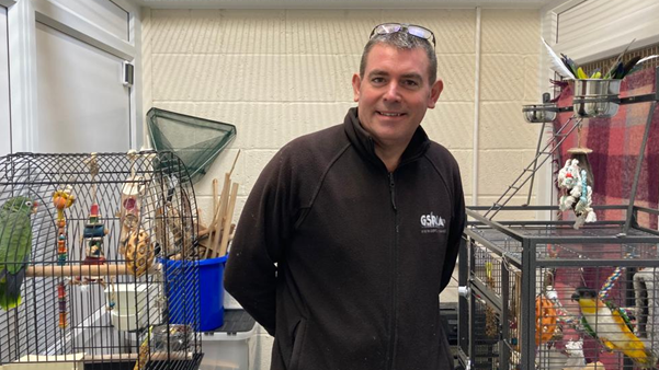 Steve  wearing a black fleece with the GSPCA logo and a pair of glasses on his head, standing in front of bird cages