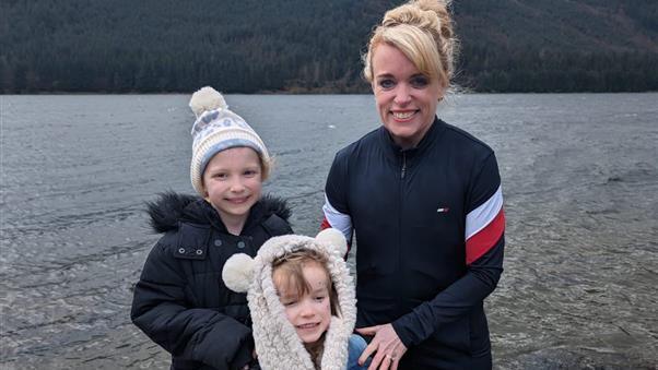 Loretta Davies has blonde hair (picture back right) and smiles at the camera with her two children stood next to her in front of a lake. Both children wear fluffy hats. 