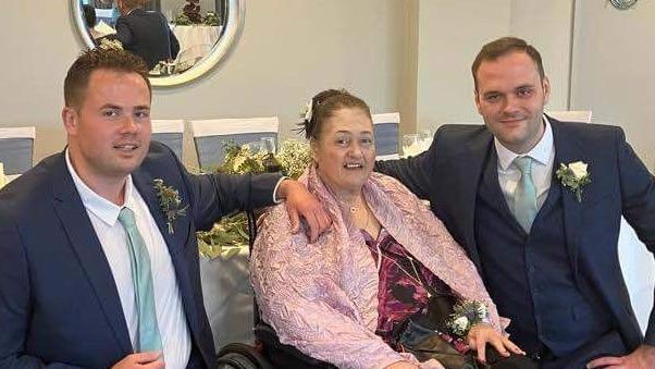 Two men in suits smiling next to an older woman in a wheelchair