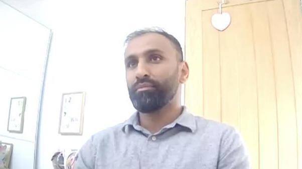 A still from a video call with a man in an office setting. He has dark hair and a beard and is wearing a grey polo shirt.