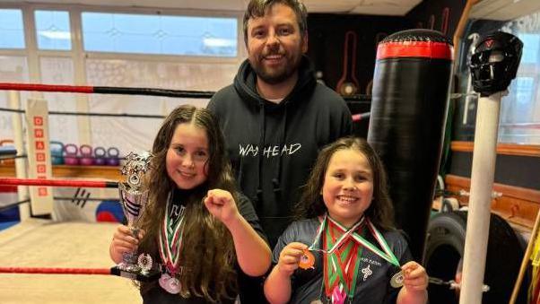 Annarose and Lili-Rose with their coach Alex Williams at their martial arts club, with all three smiling, and the two girls showing medals and a trophy