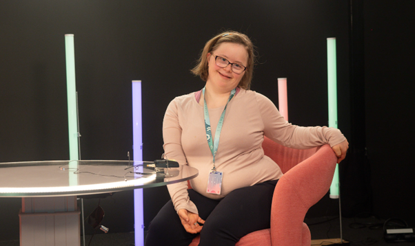 A picture of Emily Ann Riedel. She is in a TV studio. She is wearing lanyard around her neck. She has a cream coloured long sleeve top on and black leggings. She has glasses on and is smiling directly at the camera. She is sitting on a peach coloured chair, which has a circular glass table next to it. There are four neon coloured strip lights behind. The wall behind her is black.