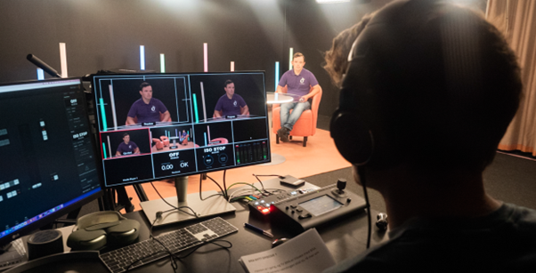 A photo a TV news studio. There is a person sitting at a computer screen with headphones on. They are looking at a man who is sitting down while he is being filmed. The same man in the background also appears on the computer screen.