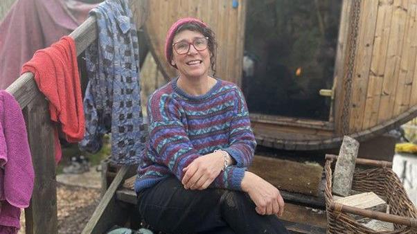 Stanmer Sauna Garden CIC director Bella Kirkus pictured sitting on steps leading into the sauna at Stanmer Park. Bella is wearing a woolly purple, red, green and blue jumper and a red hat and glasses. Behind Bella the entrance into the sauna is visible. 