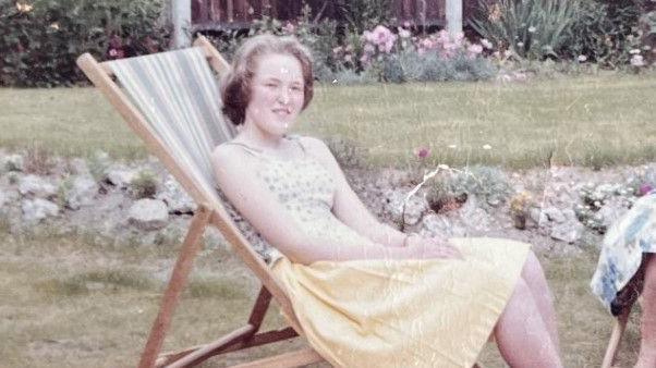 Jan Doyle, age 16, is sat in a deck chair in a garden. The photo is taken from the 60s, is grainy and she is wearing a typical 60s dress. 