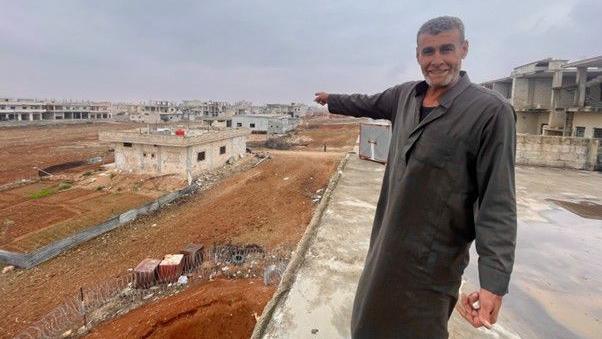 A man points into the distance at a plot of land which looks empty except for some dilapidated buildings 