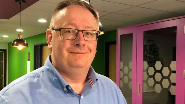 A man is wearing a blue shirt and glasses. He is in a modern office space nad smiling at the camera 