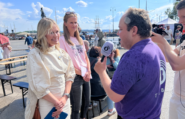 A TV BRA journalist interviews two women. He is wearing a purple t-shirt and holding a microphone, which has a white microphone cover. One woman is wearing a cream coloured top and smiling at the reporter. The other woman is wearing a pink t-shirt and listening to the interview. The interview is taking place on a sunny day. The sky is blue with a few clouds in the sky. In the right hand corner of the image a camera man stands holding a small hand held camera and has a pair of earphones on.