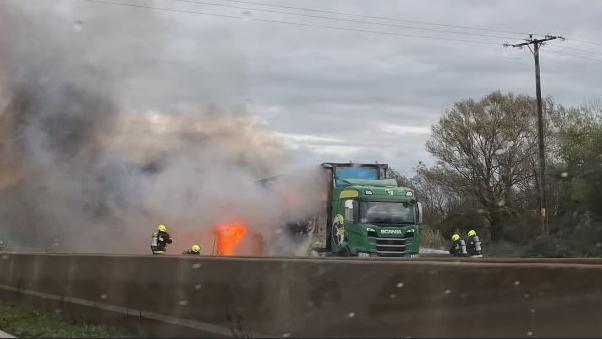 Four firefighters, in protective clothing and yellow helmets, trying to extinguish a fire on the M5. At the bottom of the picture is the motorway barrier, and above is a green lorry with flames coming out of the side. 