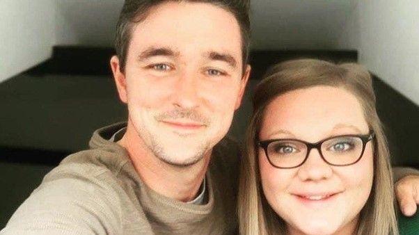 Tyler Byrne, a young man with dark hair and a short beard, standing on the left of the picture next to a woman with light brown long hair and glasses. Both of them are looking at the camera and smiling