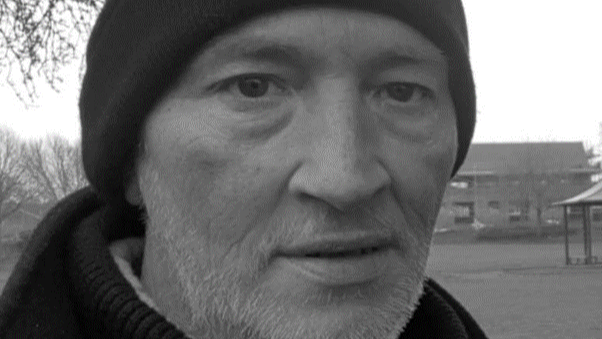 A black and white image of a man, with a short white beard and a black woolly hat.