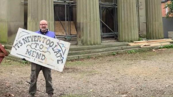 A man wearing a purple jacket and brown trousers is holding a large sign saying "it's never too late to mend". He's standing next to a rusting red skip, with a large pillared building behind him signed 'Spilsby Theatre'.