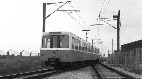 Black and white photograph, dating from 1980, showing the front of a Tyne and Wear Metro train.  It is on a track, with high wire fences on either side.