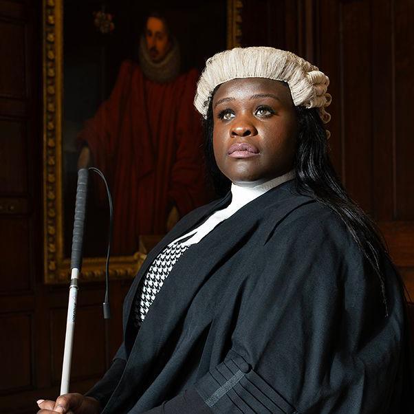 A barrister with long dark hair in her black gown and white wig. She is holding a cane and is sat with a wooden wall behind her, a serious expression on her face.
