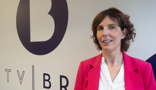 TV BRA boss Camilla Kvalhei. She is pictured in a posed photograph in front of a the TV station's logo. The wall behind her is white and the text for the company is written in black. She is wearing a pink suit style jacket and a white shirt. She has brown hair and is looking at the camera.