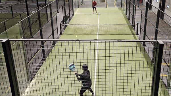 Two players inside the black mesh cage of a padel tennis court. There is a net between them and the surface is an astro turf green with a white line running down the centre. 