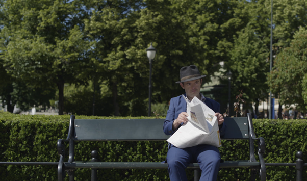 Svein Andre Hofsø sitting down on a park bench. He is wearing a navy suit and has a broadsheet newspaper partly opened on his lap. He is wearing a hat on his head appears to be looking to his left. There are trees and bushes behind him