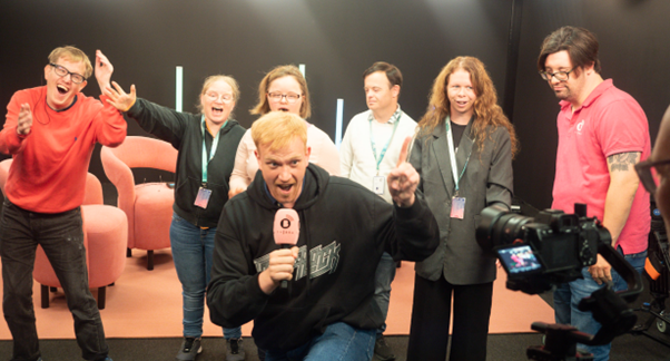 Seven reporters in the TV studio. One is in the foreground in a black hoodie and jeans. He is holding a microphone and is speaking into a camera directly in front of him He has blonde hair and is pointing the index finger of his left hand in the air. His colleagues are standing behind him and they look happy. 