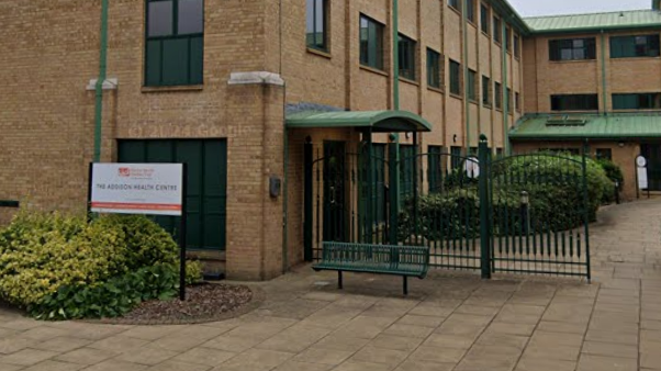 The entrance to Addison House Surgery. It is a building of several levels made out of cream bricks. There are green gates and a bench out the front, as well as a white sign advertising its name.