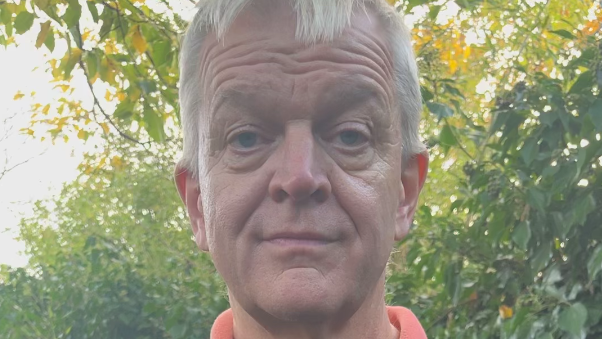 A middle-aged man with white hair. He is stood in front of a green hedge looking into the camera