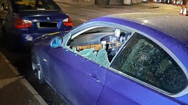 A dark purple car with a smashed passenger window. The car is parked along the side of the street, behind a dark blue BMW. 