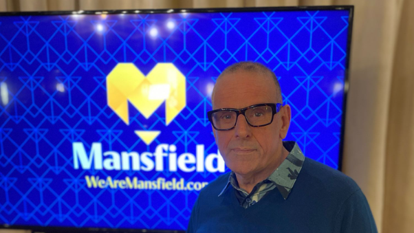 A man in glasses stands in front of a sign for Mansfield