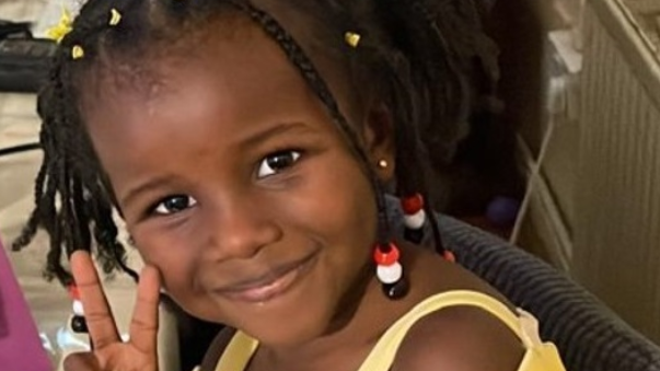 A four-year-old girl in a yellow dress holding her fingers in a peace sign and smiling