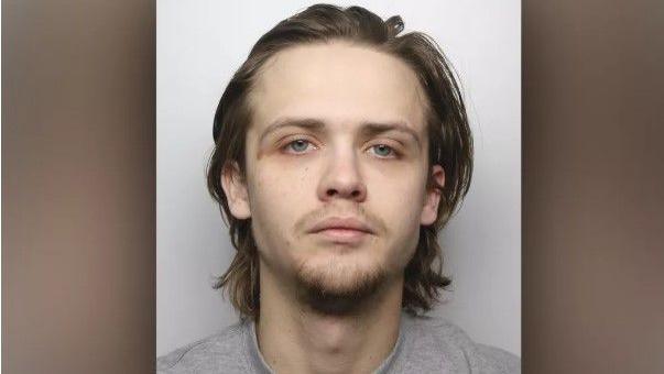 A police mugshot of Jack Naylor, who has long brown hair and a whispy brown goatee. He stares into the camera with a blank expression.