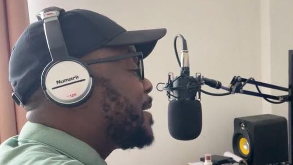 A man speaks into a microphone with silver headphones on. 