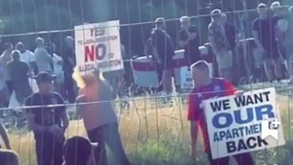 Protesters are seen through a fence holding signs which read "Yes to legal immigration, no to illegal immigration" and "We want our apartments back"