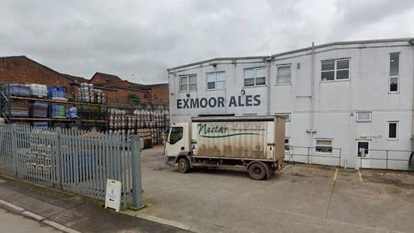 A photo of the factory in Wiviliscombe from the outside, a white two storey building with barrels of beer stacked up outside and a truck parked in front.