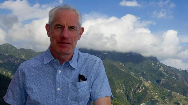 Tudor Evans, a man in his 60s with grey hair, smiling for a photo in front of mountains. He is wearing a short-sleeved blue shirt and his sunglasses are tucked into his shirt pocket