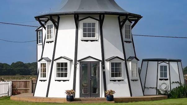 A white and black curved two-storey house with wooden decking outside and green grass