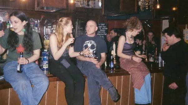 Three woman and two men sat on a bar talking and drinking. In the background there are beer pumps and more people.