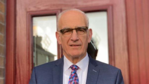 The Rev John Greer - a bald man looking to camera and wearing glasses, a dark blue blazer, white shirt and multi-coloured tie