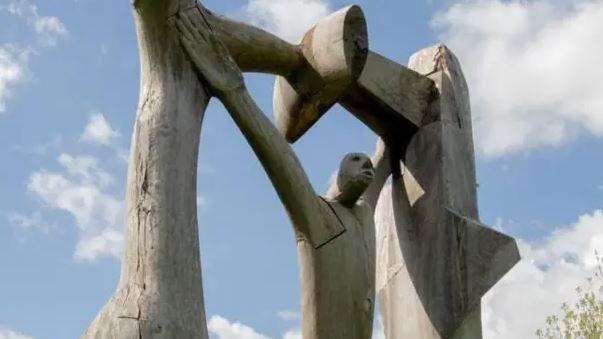 A wooden sculpture of a man with his arms in the air, resting them against a wooden frame. There is a blue sky with white clouds.