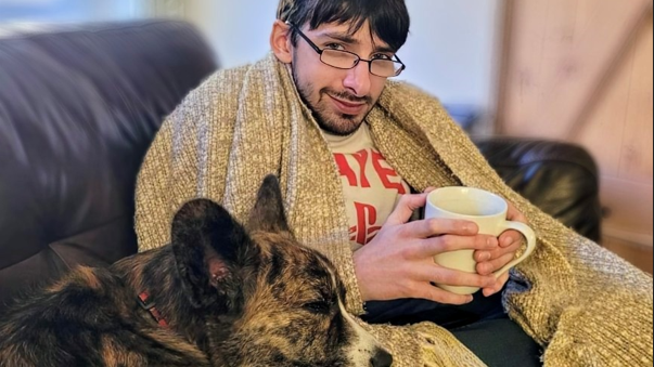 Daniel is sat on a settee with a blanket around him and a dog resting on his lap. He is holding a mug. He has glasses and a short beard.