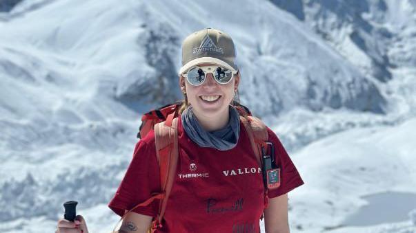 Adriana Brownlee stands and smiles as she climbs a mountain, wearing a red t-shirt and backpack, brown hat and polarised sunglasses