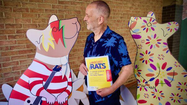 Rick Welton holding a sheet of paper that says "Mischief Rats" on it, flanked by two large rat artworks either side of him. One has a red and white stripy top holding a guitar with face paint and the other is yellow with flowers