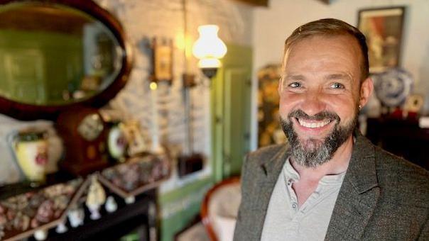 Dave is standing inside the room with a wooden-framed mirror. He has brown hair and a beard, with strands of grey in it. He is wearing a tweed coast over a light brown shirt. He is smiling as he looks into the camera.