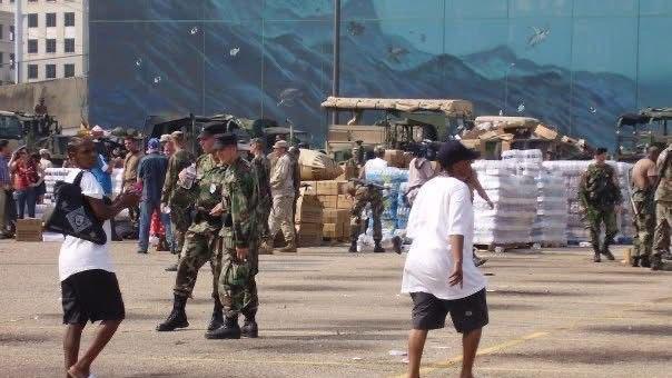 Soliders stand with guns in front of boxes as people wearing white tops and black shorts walk towards the boxes.