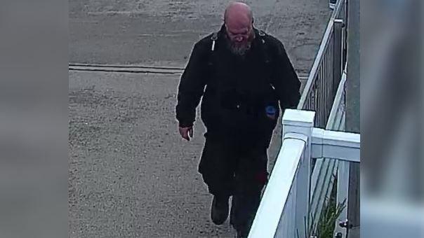 A man wearing a dark green bomber jacket, black cargo trousers and a black backpack. He is bald with a beard and is walking with his head facing the ground. There is a white fence to the right of the image. 